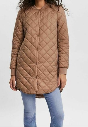 Vrouw die een lange, lichtbruine doorgestikte jas met drukknopen, geribbelde manchetten en lichtblauwe jeans draagt, tegen een neutrale achtergrond.