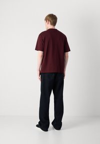 Burgundy cotton t-shirt with short sleeves, relaxed fit, plain design. Worn with dark denim pants, additional details not visible.