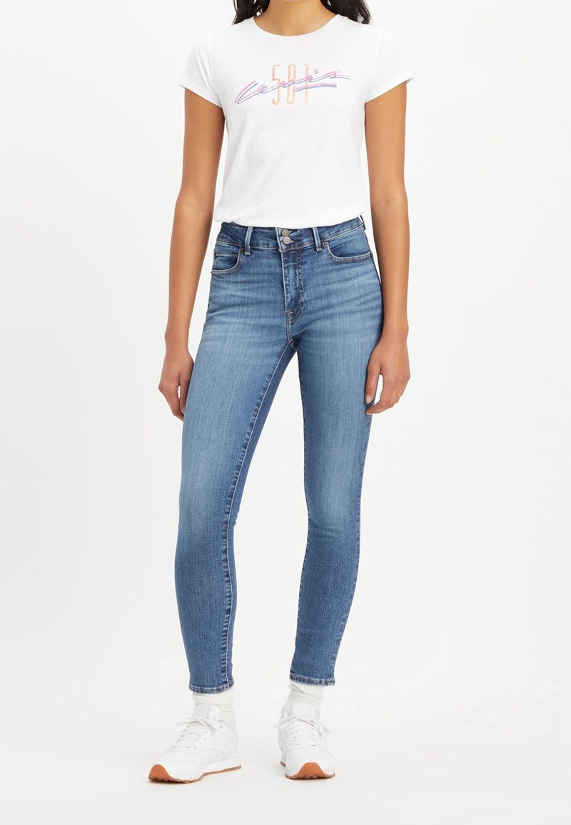 T-shirt blanc à manches courtes avec un graphique multicolore "501", associé à un jean skinny bleu clair. Le mannequin porte des baskets blanches avec des accents orange.