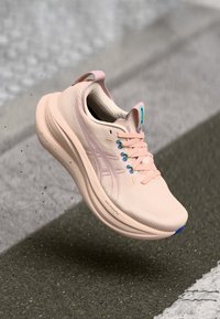 Chaussure de course rose clair avec une tige texturée, des accents bleus et des lacets contrastants. Elle possède une semelle rembourrée pour le confort et le soutien.