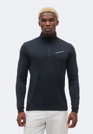 Mann mit kurzen blonden Haaren, der ein schwarzes Langarmshirt mit Viertelreißverschluss und weiße Hose trägt, blickt nach vorne vor schlichtem Hintergrund.