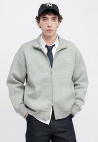 Sweatshirt cinzento claro com fecho zip, feito de material polar, apresentando dois bolsos frontais, punhos canelados e um pequeno logotipo no peito.