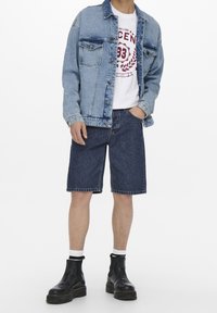 Denim-takki vaalealla pesulla, valkoinen t-paita, jossa on punainen grafiikka, tummat denim-shortsit ja mustat nilkkurit paksuilla pohjilla.