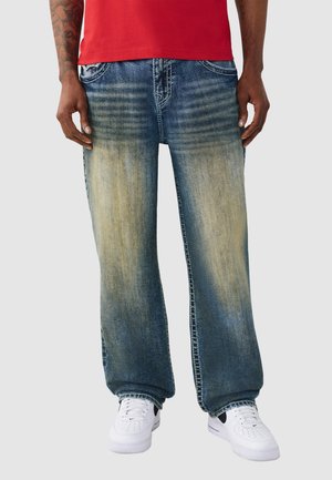 Person, die ausgewaschene blaue Jeans mit weitem Bein trägt, kombiniert mit weißen Sneakers und einem roten Hemd, steht vor einem schlichten Hintergrund.