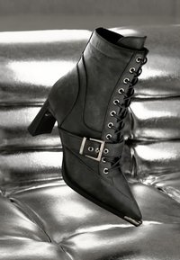 Schwarze Leder-Ankelstiefel mit spitzem Zehenbereich, Schnürung an der Vorderseite und einem Seitenriemen mit einer silbernen Schnalle. Stiletto-Absatz und strukturierte Oberfläche.