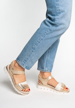 Feet wearing beige and white open-toe sandals with thick white soles, paired with light blue rolled-up jeans on a white background.