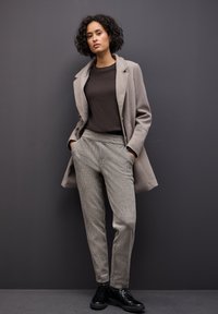 Lichtgrijze jas, donkerbruine trui en gestructureerde grijze broek met een patroon. Zwarte veterschoenen. Neutrale achtergrond.