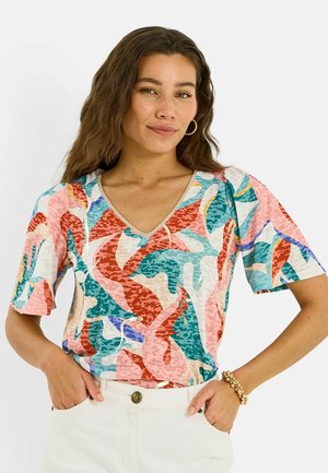 Mujer con cabello largo y ondulado que lleva grandes pendientes de aro, una camisa con cuello en V de patrón abstracto y colorido, pantalones claros y una pulsera de cuentas.