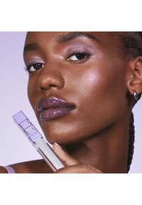 Glanzend paarse lippenproduct vastgehouden naast het gezicht van een model. Het model heeft glinsterende oogschaduw en highlights, met een egale huidtextuur.