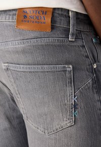 Jeans din denim gri, cu un eticheta din piele maro cu text albastru. Cusături decorative notabile în albastru și verde pe buzunarul din spate.