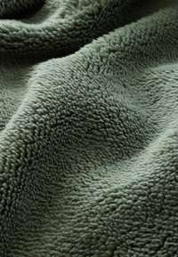 Miękka, teksturowana koc fleece w przygaszonym zielonym kolorze. Tkanina ma pluszowy, gruby włos z lekko połyskującą powierzchnią, co nadaje jej przytulny wygląd.