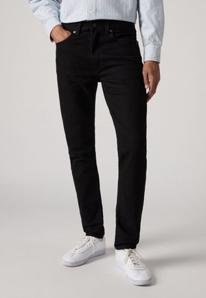 Homme portant un jean noir slim, des baskets blanches et une chemise à manches longues rayée bleue et blanche, debout avec une main dans la poche.