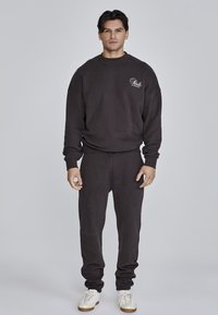 Felpa grigia scura e pantaloni jogger realizzati in morbido tessuto. Collo rotondo, vestibilità ampia, polsini a coste. Logo bianco sul petto. Sneakers bianche.