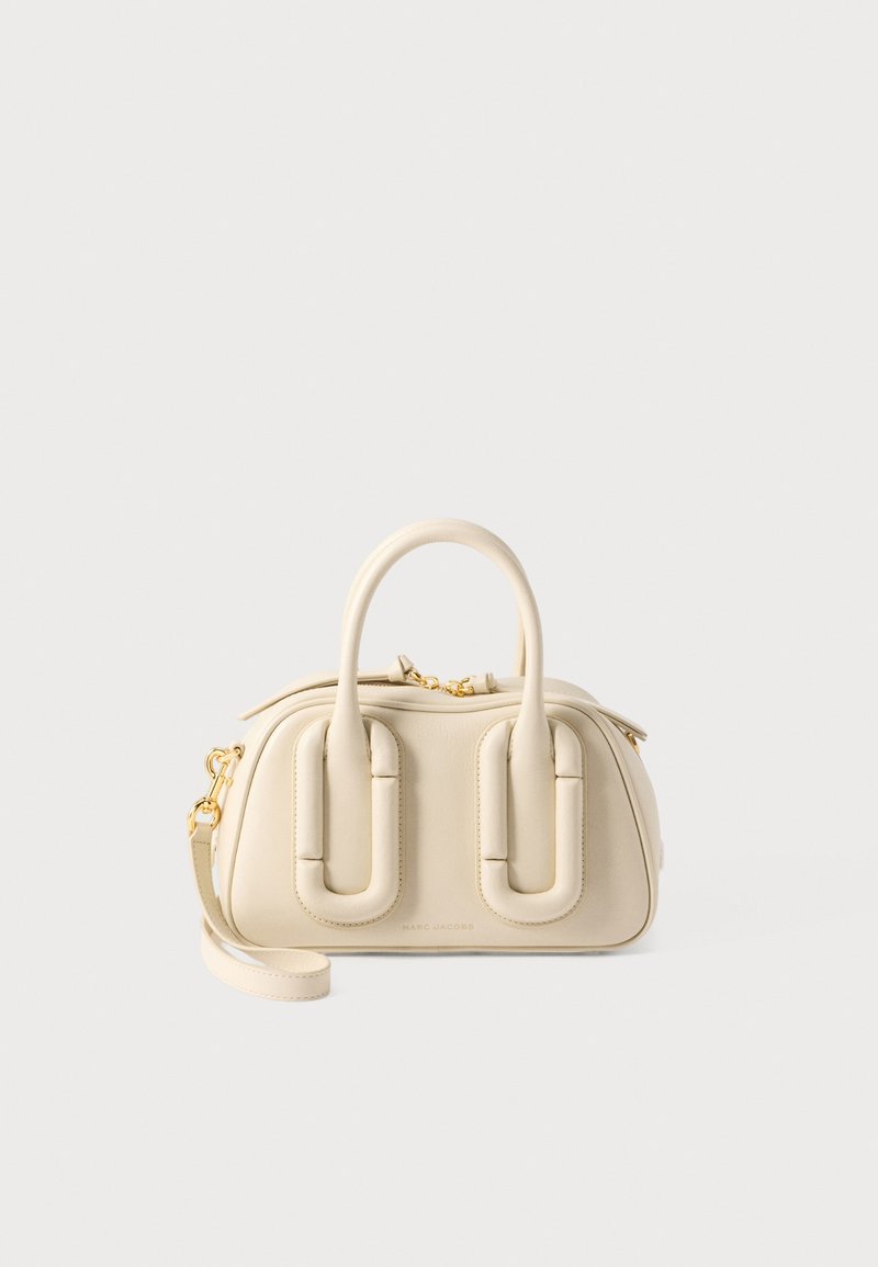 Petit sac à main de couleur crème avec des poignées arrondies, une fermeture éclair et un fermoir dorés, et une bandoulière amovible sur un fond clair uni.