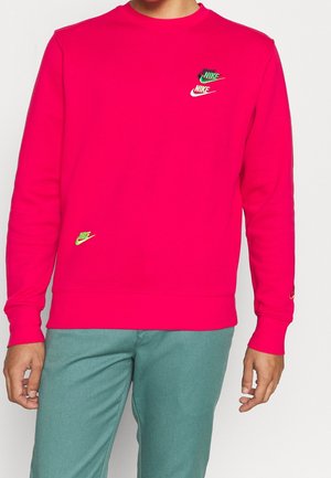 Osoba nosząca jasnoróżową bluzę z małymi, wielokolorowymi logo Nike i zielone spodnie na białym, jednolitym tle.