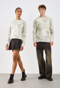 Dwa modele noszą długie rękawy, swetry tie-dye w stonowanej zieleni i beżu. Modelka ma na sobie plisowaną czarną spódnicę z denimu i czarne buty wojskowe. Model męski nosi luźne, ciemne dżinsy.