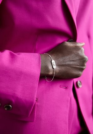Bracelet en argent avec un charme rectangulaire, porté sur une main à la peau foncée. L'arrière-plan présente un blazer rose vif avec un tissu texturé.