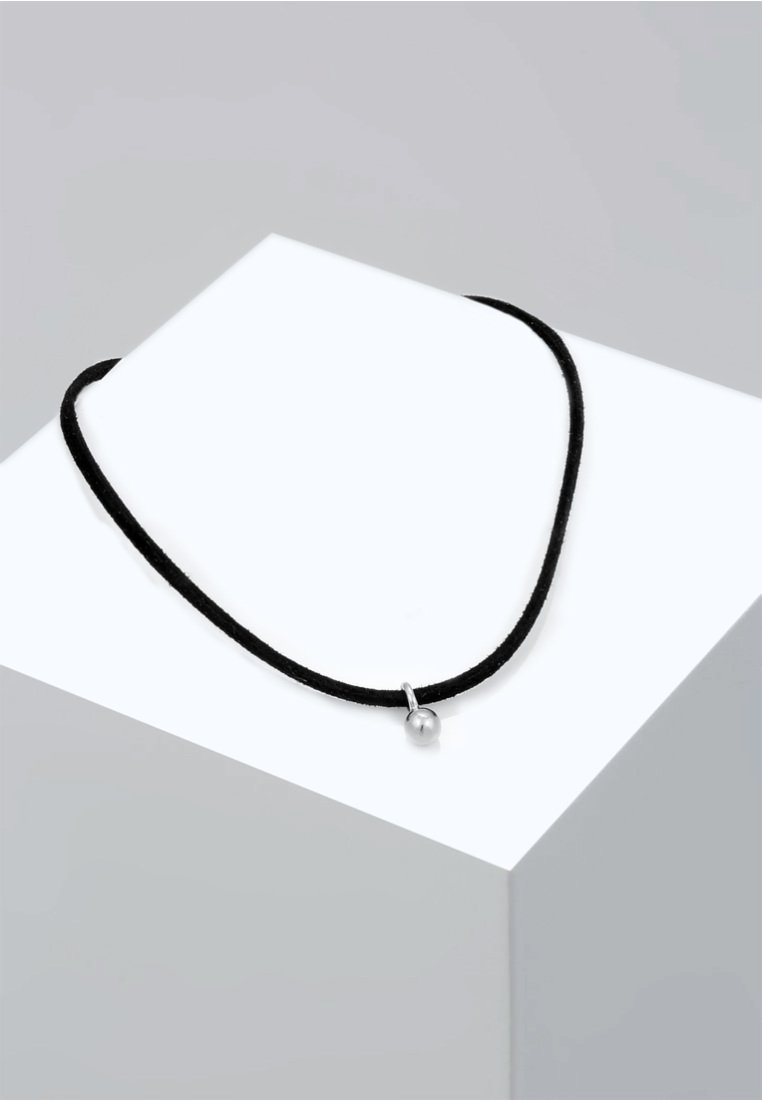 Schwarzes Schnur-Halsband mit einem silbernen Pendant in Form einer Kugel, das eine glatte Textur und ein einfaches Design aufweist, auf einem weißen geometrischen Display ruht.