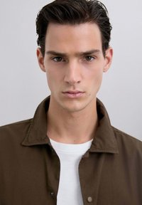 Tânăr cu părul scurt și închis la culoare, purtând o jachetă maro peste o cămașă albă, privind înainte cu o expresie neutră, pe un fundal simplu.