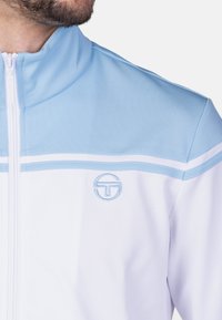 Weißer Trainingsjacke mit hellblauen Akzenten, hohem Kragen, Reißverschluss und gesticktem Logo auf der linken Brust. Strukturiertes Material.