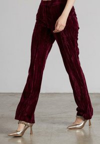 Persona con pantalones de terciopelo burdeos texturizados y tacones altos metálicos dorados con correas de tobillo joyadas, de pie sobre un suelo de hormigón.