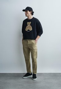 Homme portant une casquette noire, un sweat-shirt noir avec un dessin d'ours en peluche, un pantalon cargo beige et des baskets noires, debout contre un mur gris uni.