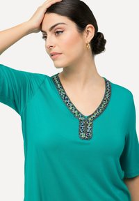 T-shirt a scollo a V in teal, con decorazioni in perline blu e verdi attorno al collo. Maniche corte e texture di tessuto liscia.