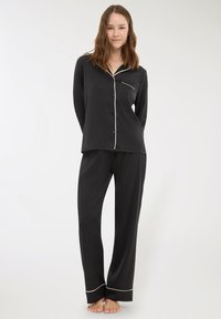 Giovane donna in piedi a piedi nudi, indossa un completo pigiama nero con bottoni e piping bianco, su uno sfondo chiaro e semplice.