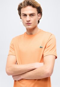 Νέος άνδρας με ανοιχτό καστανά μαλλιά που φοράει ροζ κοντομάνικη μπλούζα με μικρό πράσινο λογότυπο κροκόδειλου, με τα χέρια σταυρωμένα, σε απλό φόντο.