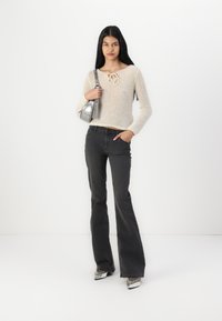 Strikket sweater i creme med en dekorativ snørelukning ved halsen, parret med sorte flagrende jeans og sølvfarvede hæle. Accessories omfatter en sølvfarvet håndtaske.