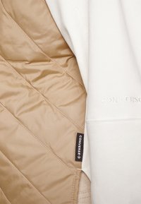 Interior forrado de lana blanca con patrón acolchado en marrón claro y etiqueta marrón contrastante que muestra "CONVERSE". Textura suave, material cálido.