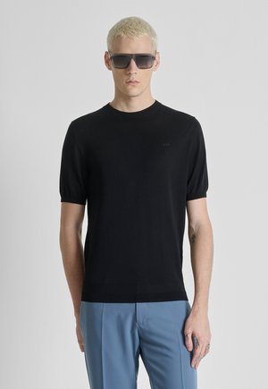 Persona rubia que lleva grandes gafas de sol negras, una camiseta negra de manga corta y pantalones azul claro, posando frente a un fondo liso y claro.