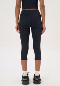Zwarte capri leggings gemaakt van rekbaar materiaal, met een hoge tailleband, een gladde textuur en reikt tot halverwege het onderbeen, gecombineerd met zwarte sneakers.