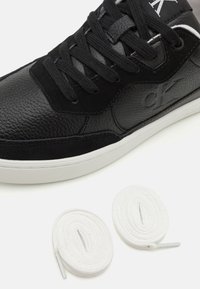 Sneaker in pelle nera con superficie texturizzata, dettagli in suede e lacci neri piatti. Include due lacci bianchi mostrati accanto alla scarpa.