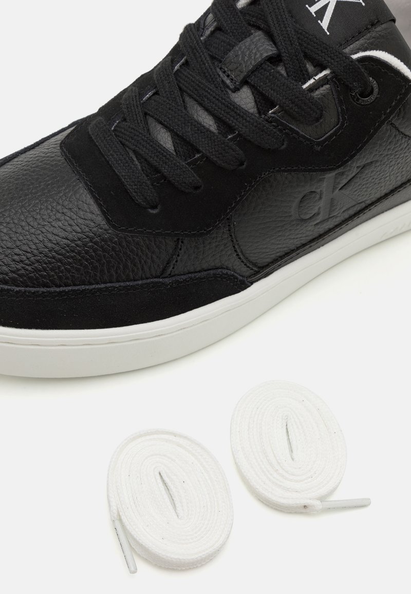 Baskets en cuir noir avec surface texturée, accents en daim et lacets noirs plats. Comprend deux lacets blancs affichés à côté de la chaussure.