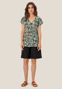 Kortärmad, V-ringad topp i ljusblått med svart och brunt blommönster; matchad med svarta shorts och bruna sandaler.