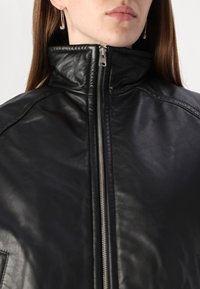 Veste en cuir noir avec col haut, dotée d'une fermeture éclair argentée et d'une texture lisse. Le design inclut des détails cousus aux épaules et une poche devant.