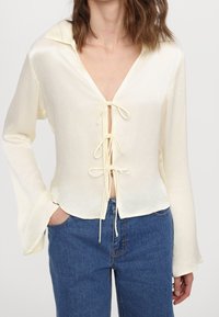 Løs, cremefarvet bluse med dyb V-udskæring og bånd foran. Lange ærmer i klokkeform og let tekstureret stof.