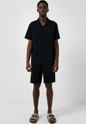 Mężczyzna stojący w czarnej, teksturowanej koszuli na krótki rękaw z guzikami oraz dopasowanych szortach, noszący beżowe sandały na klamry na jednolitym tle.