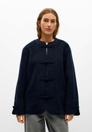 Jeune personne aux cheveux châtains clairs portant une veste bleu marine foncé boutonnée et un pantalon gris, debout devant un fond blanc uni.
