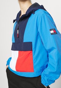 Color-blocked jacket in blue, navy, red, and white. Features a zippered neck, drawstring hood, and logo patch on the sleeve. Lightweight fabric.