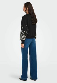 Črna majica s kapuco z rožastim vezenjem na rokavih, sproščen kroj, v kombinaciji z modrimi širokimi hlačami, prikazana s hrbtne strani.