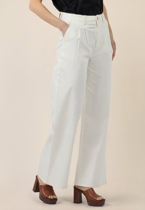 Vrouw draagt een witte, wijde, hooggetailleerde broek met zakken, gecombineerd met bruine platform sandalen met open tenen en een ingestoken zwarte top.