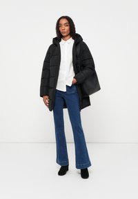 Schwarze gepolsterte Jacke mit Kapuze, weißes Hemd mit Knöpfen, weit ausgestellte blaue Jeans und schwarze Stiefeletten. Enthält eine schwarze Umhängetasche.