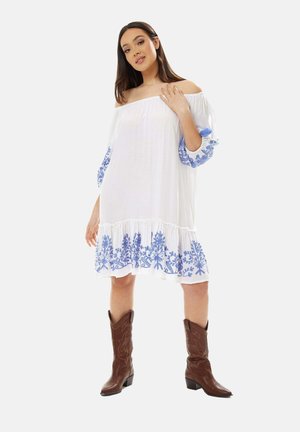 White off-the-shoulder dress with blue floral embroidery at sleeves and hem, featuring a tiered ruffle design. Brown cowboy boots complete the look.