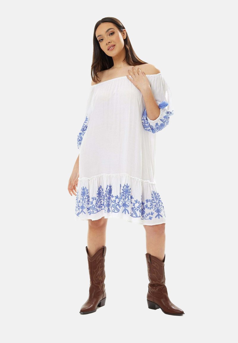 White off-the-shoulder dress with blue floral embroidery at sleeves and hem, featuring a tiered ruffle design. Brown cowboy boots complete the look.