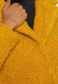 Cappotto giallo con una superficie trama morbida e tattile. Collo ripiegato e dettagli di cucitura visibili. Tessuto nero sotto il collo.