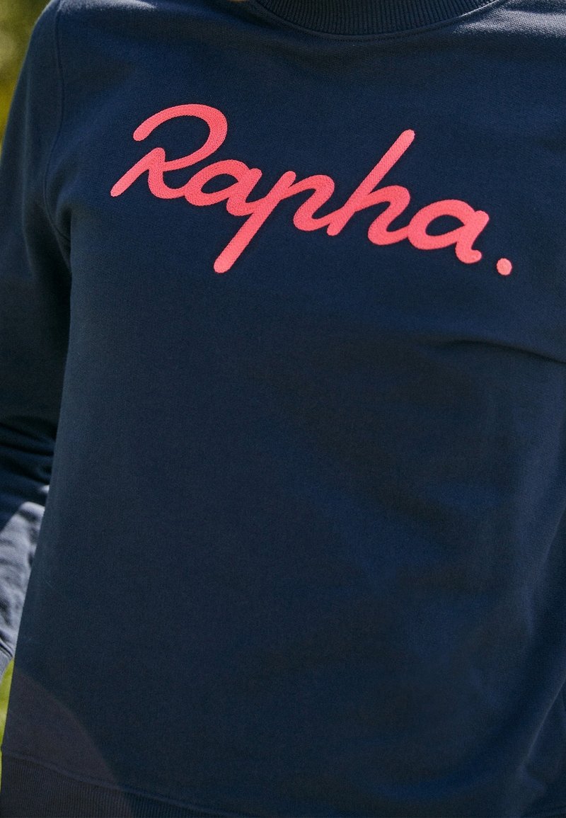 Sweat-shirt bleu foncé avec le mot "Rapha." en grande écriture rose sur la poitrine.