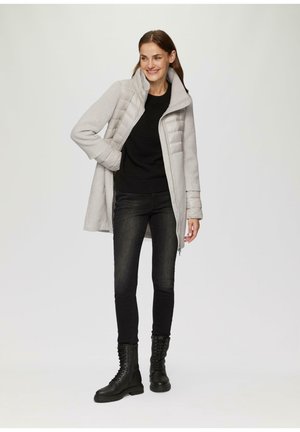 Grijze hybride jas met een gewatteerde voorkant, hoge kraag en rits, gecombineerd met een zwarte structuurtrui, donkere jeans en zwarte veterschoenen.