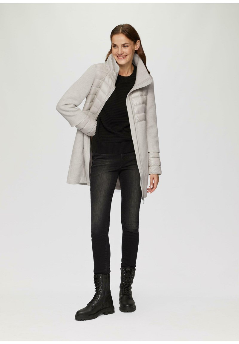 Grauer Hybridmantel mit gepolstertem Vorderteil, hohem Kragen und Reißverschluss, kombiniert mit einem schwarzen strukturierten Pullover, dunklen Jeans und schnürbaren schwarzen Stiefeln.